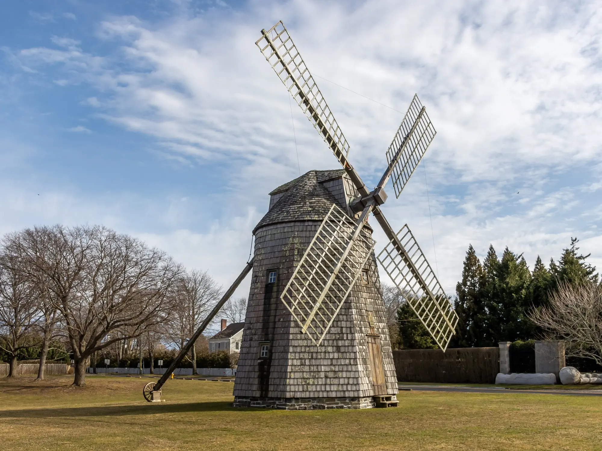 A shingled windmill on a lawn under a grey March sky, with bare trees behind